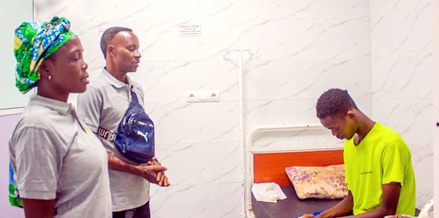 Two volunteers pray with a young man seated on his hospital bed